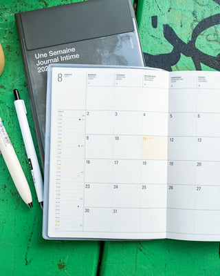 An open planner calendar layout on white pages, placed on a green wooden table next to a closed dark gray journal and two white pens.