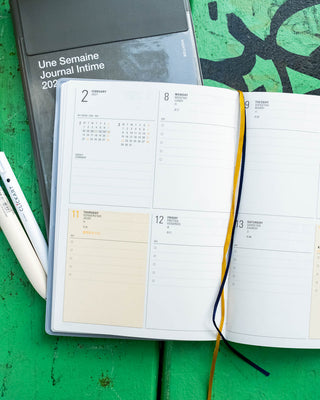 An open weekly planner on a textured green wooden surface, next to a closed copy of the same planner and two white pens.