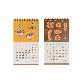 spiral-bound desk calendars shown side by side on a plain white background. The left calendar has an orange/yellow top section, the right has animal shaped bread on a brown background