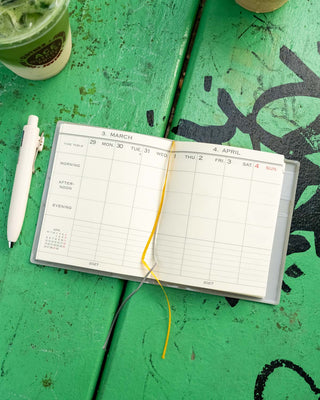 A weekly planner white paper pages laid open on a green wooden surface. The planner has yellow and gray ribbon bookmark