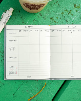 An open weekly planner spread on a green tabletop, with a white pen 