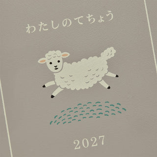 A close-up of a planner cover with a cute illustration of a white fluffy sheep jumping over small green hills, with Japanese text at the top and the year 2027 at the bottom.
