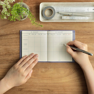 An open weekly planner sits on a wooden desk, with a person writing. A small plant and a tray stationery items are nearby
