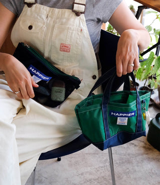Person holding a green 'Happier' bag with happier label layer pouch bag in their lap, sitting by plants