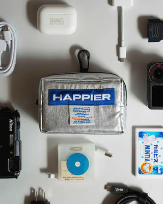 Collection of items including a 'Happier' bag, cameras, and electronic devices on a white surface.
