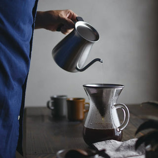 Person pouring water from a kettle into a coffee pour over carafe on a wooden table.