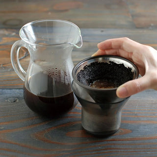 Hand holding a coffee filter with ground coffee next to a glass carafe on a wooden table