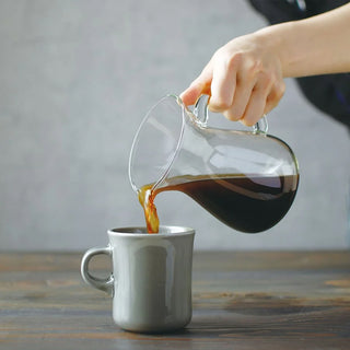 Person pouring coffee from a glass carafe into a gray mug on a wooden surface.