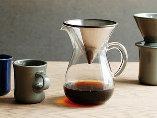 A clear glass pour-over coffee carafe with a metal cone filter, filled with coffee, on a table with mugs