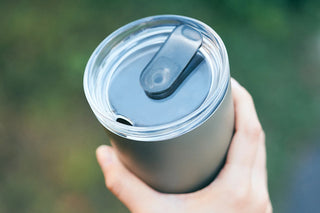 Hand holding a silver tumbler with a clear lid against a blurred green background