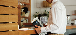 Person standing at coffee counter with newspaper and notebook in hand, sticky notes clipped onto notebook