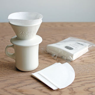 White ceramic mug with ceramic brewer and paper filter inside next to packaging of coffee filters on a wooden table