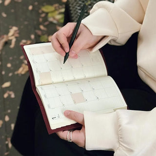 Person holding a notebook calendar and pen, with a blurred outdoor background