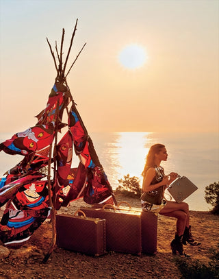 Person sitting by a lake with a colorful fabric and suitcase at sunset.