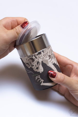 Tea canister held in hands; silver lid lifted, showing opening and skeleton print