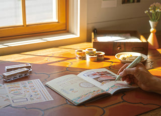 Person using a notebook with stationery items on a wooden desk near a window.