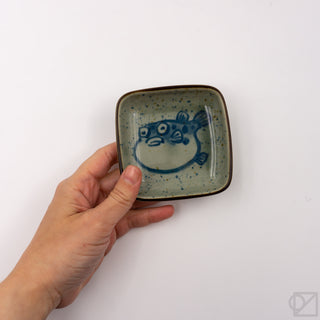 Hand holding small square ceramic puffer-fish dish with blue fish graphic on speckled glaze