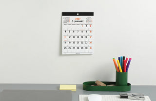 A minimalist 2027 desk calendar hanging on a light gray wall above a desk with stationery, including a green pen cup, yellow sticky notes
