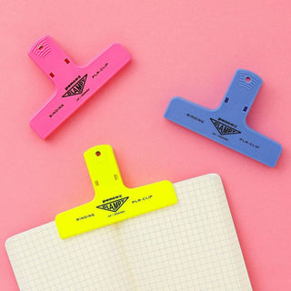 Neon Yellow, Light Blue, and Pink penco clips on a pink background. The Yellow binder clip is holding a notebook open.