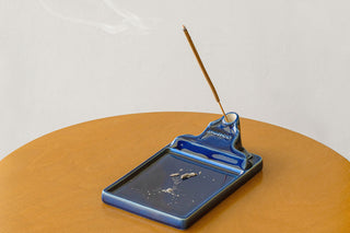 A blue ceramic incense holder with a single burning incense stick, set on a wooden table