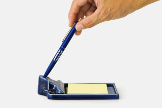 A hand holding a blue pen over a blue ceramic tray with a yellow sticky note pad, on a light gray background.