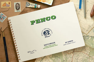 A Penco sketchbook shown on a wooden desk surrounded by vintage maps, pencils, and art supplies. 