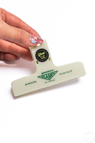 A hand holding a glow in the dark PENCO Binding Clip laid on a white background.
