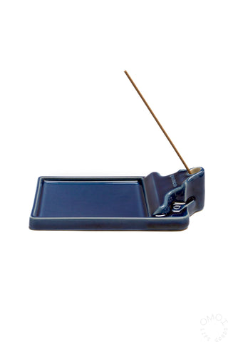 A dark blue ceramic incense holder with a single incense stick inserted, shown against a white background.