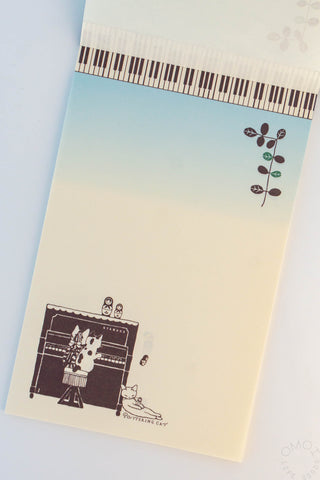Memo pad with a gradient cover from blue to beige, featuring illustrations of cats around a piano and a Matryoshka doll falling
