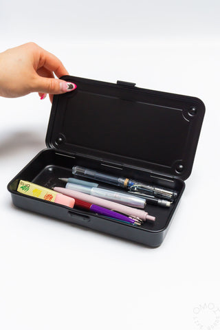 Black trunk toolbox open with pens and pencils inside; hand holding lid.