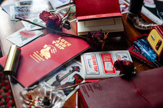 LOVE AND TRIP red notebooks a mixed in on a messy tabletop strewn with red items that match it, like red cassette tapes, dried roses, playing cards, a heart shaped ashtray, etc.