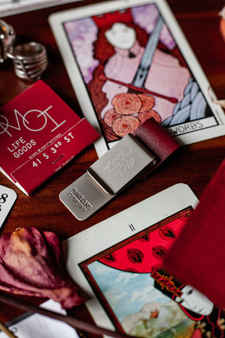 A matching metal pen clip, etched with "LOVE AND TRIP" logo. It's surrounded by various items that compliment its deep red burgundy color, like matchbook, playing cards, and dried roses.