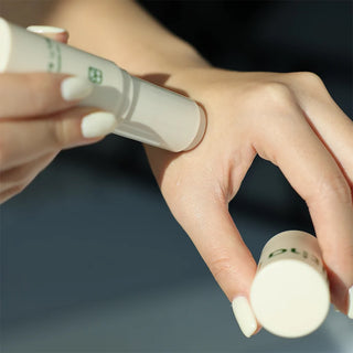 Hand applying balm stick to skin; beige tube with green logo visible