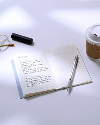 An open notebook with handwritten notes on a white surface, with a silver mechanical pencil, eyeglasses, and a coffee cup.