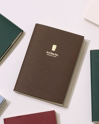 A dark brown notebook with gold foil text reading 'Tea & Diary Time - Morning Diary' surrounded by other notebooks on a light background
