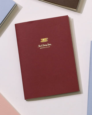 A burgundy notebook with gold foil text reading 'Tea & Time' and 'WEEKEND DIARY' on a white surface with partial notebooks visible at the edges.