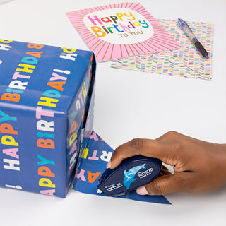 Hand using a blue recycled paper tape dispenser with a 'Happy Birthday' box and card in the background.