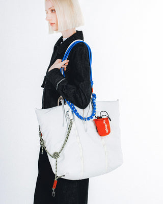 Person wearing a large white puffy tote bag. The bag features a bright blue braided shoulder strap and a small orange pouch hanging from the side 