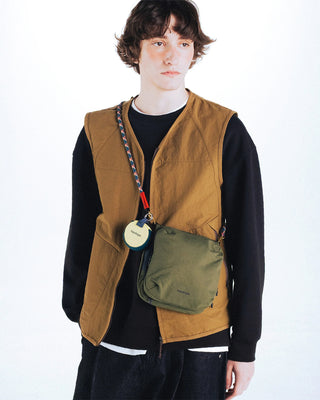 Person wearing a brown vest with a green bag over the shoulder with colorful strap on a white background