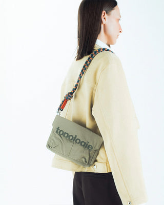 Person wearing a beige jacket with a green bag with colorful strap featuring 'topologie' branding on a white background
