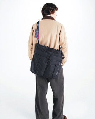Black helmet bag worn crossbody with front flap pocket and shoulder strap