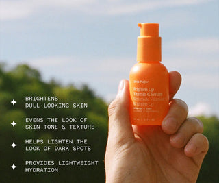 Hand holding an orange serum bottle with text about its benefits against a blurred natural background