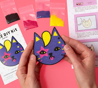 Sand art cat craft piece held by hands, with colorful sand and kit instruction sheets