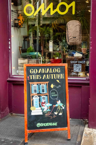 Storefront window with plants and a colorful Go Analog This Autumn sidewalk sign