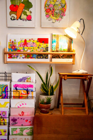 An Anglepoise lamp sits on a wooden plant stand with a few succulent plants, its light directed at a wire rack full of colorful risograph prints by local artists.