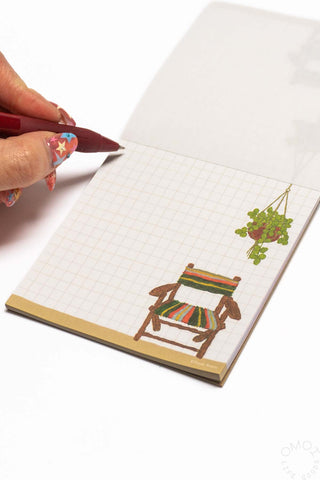 Memo pad with a page featuring a chair and plant, with a hand holding a pen