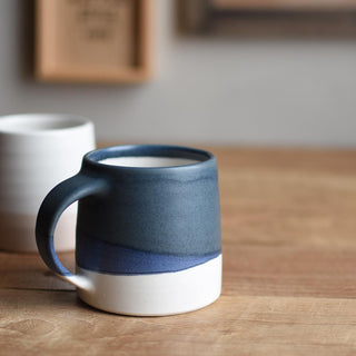Two ceramic mugs, one with a blue handle and white body, the other white, on a wooden surface.