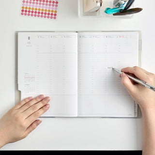 An open weekly planner spread on a white desk, with a person writing. The planner is surrounded by other stationery items on the desk