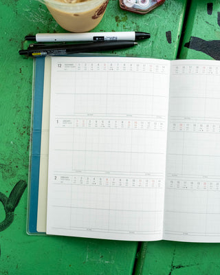 An open monthly calendar planner placed on a green wooden surface alongside pens and an iced coffee cup.
