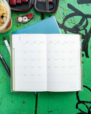 An open planner spread with blank monthly calendar pages shown on a bright green table, surrounded by stationery items including a blue notebook, pen, tape dispenser, and red stationery kit.